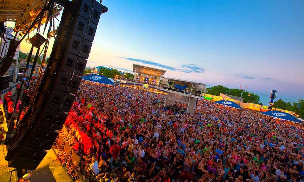 A diverse crowd of music lovers enjoying Summerfest, an outdoor music festival. Musicians performing on multiple stages, surrounded by colorful banners and decorations. People dancing, singing, and relaxing in a lively and vibrant atmosphere.